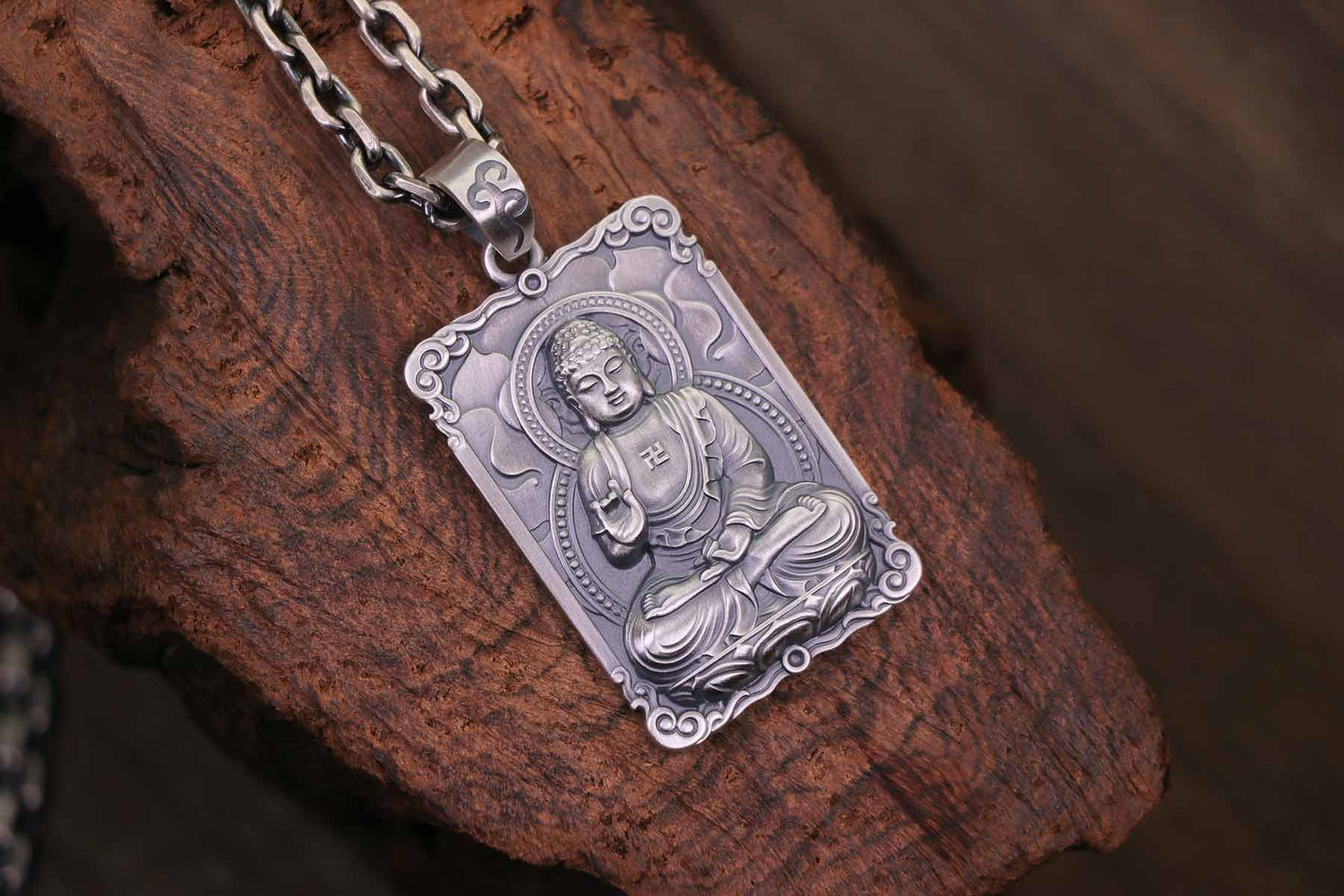 A framed silver pendant with a seated Buddha engraving and auspicious symbol, resting on wood.