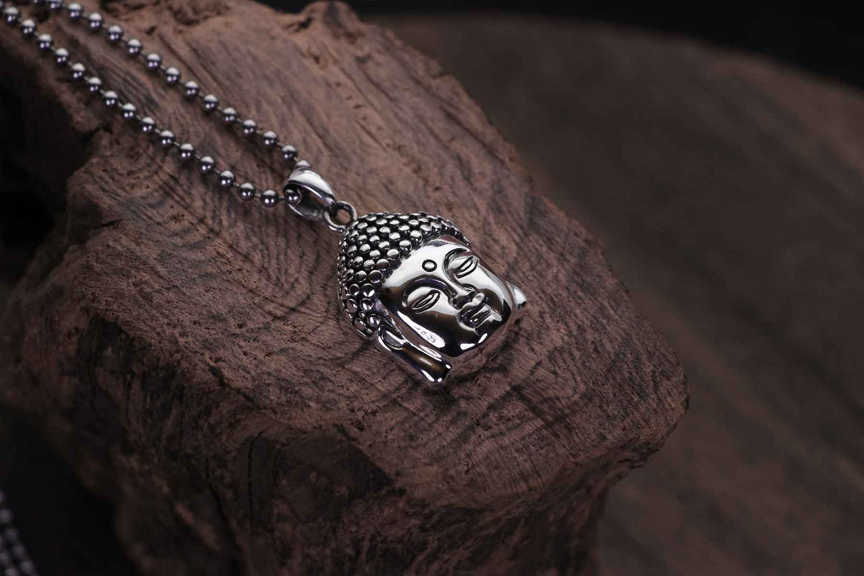 A silver Buddha head pendant resting on a piece of weathered wood.