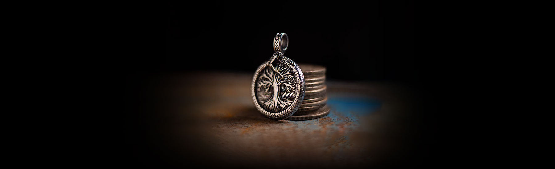 A silver Yggdrasil pendant featuring an intricate Tree of Life design, standing upright against a stack of coins on a textured surface with a dark, moody background.
