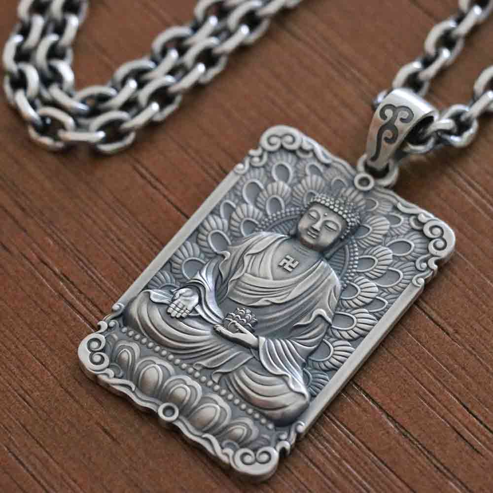 An antique silver-toned pendant necklace featuring an intricately carved Buddha figure, resting on a dark brown wooden surface with a matching chain visible in the background.